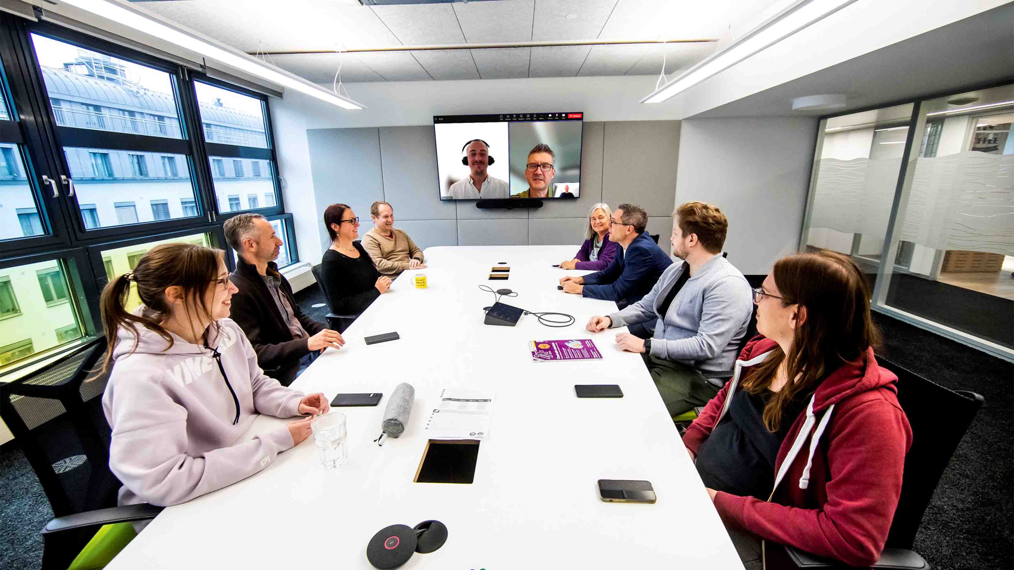 Four women and four men sit at a large meeting table in a modern room. Two other men from home offices take part in the meeting via screen.
