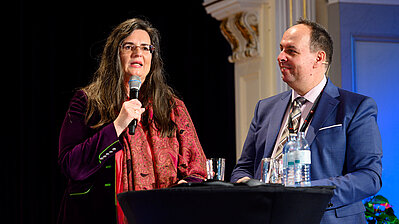 Dame mit Mikrofon und Herr in Anzug und Krawatte im Dialog auf der Bühne.