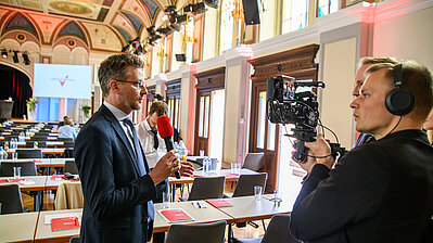 Mann in Anzug und Krawatte gibt einem Redaktionsteam ein Interview vor der Kamera.