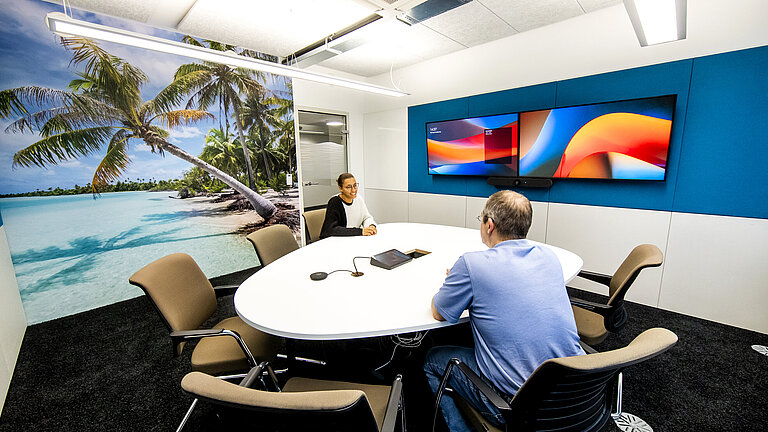 Monitor zeigt eine hybride Besprechung mit Menschen vor Ort sowie im Home Office per Video zugeschaltet.