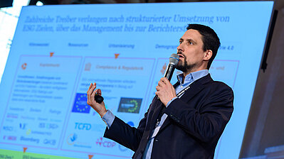 Man in suit with microphone in hand in front of a screen on stage of Casino Baden.
